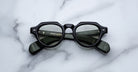 A pair of Jacques Marie Mage Kellerman thick black round eyeglasses with dark tinted lenses rests on a white marble surface, folded arms revealing text inside the frames.