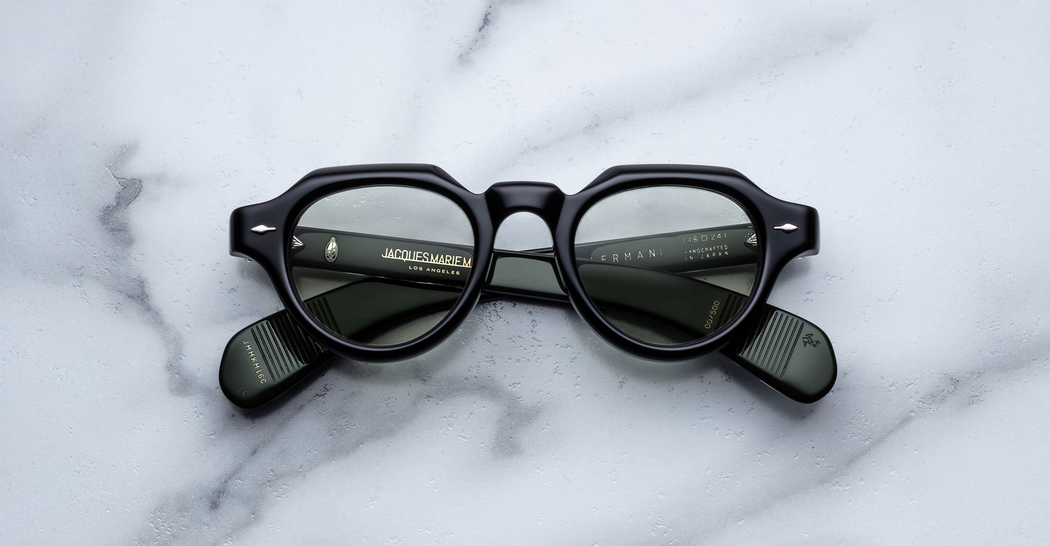 A pair of Jacques Marie Mage Kellerman thick black round eyeglasses with dark tinted lenses rests on a white marble surface, folded arms revealing text inside the frames.