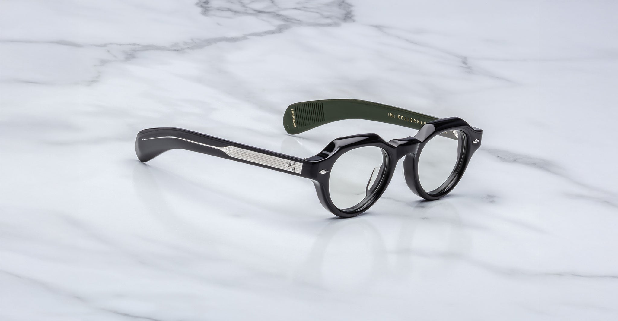 Jacques Marie Mage Kellerman black round eyeglasses with thick frames and green inner arms rest on white marble. One arm is labeled inside, while the temples display a metal accent.