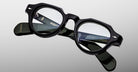 A pair of Jacques Marie Mage Kellerman black, thick-rimmed eyeglasses with round lenses is displayed on a reflective surface, revealing detailed branding and product information inside the arms.