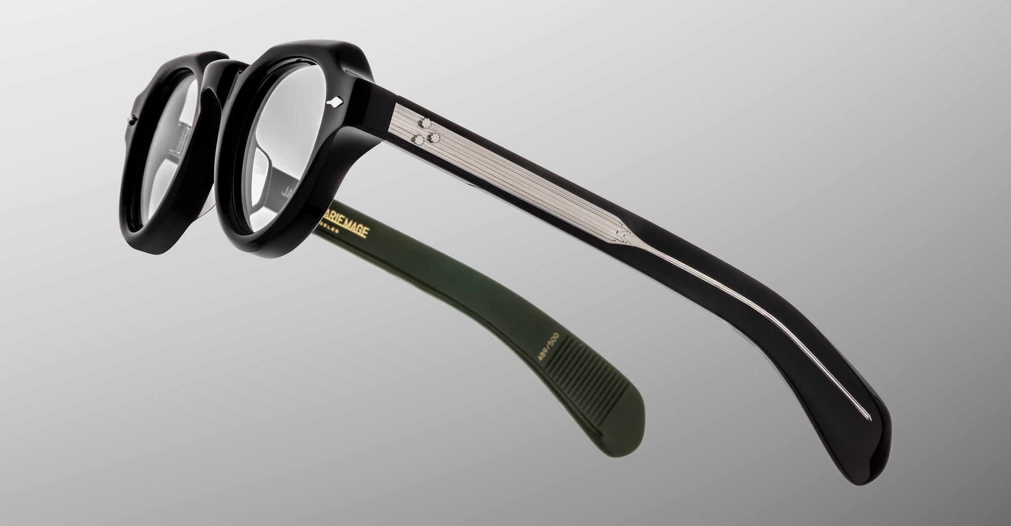 A close-up of Jacques Marie Mage Kellerman black round eyeglasses featuring silver temple detailing and one green temple tip, set against a light gray gradient background.