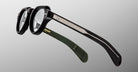 A close-up of Jacques Marie Mage Kellerman black round eyeglasses featuring silver temple detailing and one green temple tip, set against a light gray gradient background.