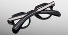The Jacques Marie Mage Kellerman eyeglasses, featuring black round frames with silver accents, are shown folded with crossed temples on a plain gray background.