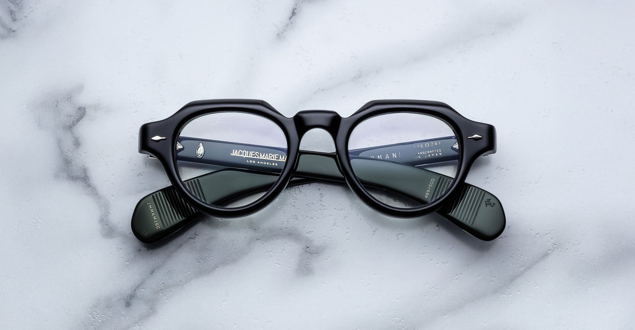 A pair of Jacques Marie Mage Kellerman black, round eyeglasses rests on a white marble surface, with brand details in white text visible on the inner arms through the clear lenses.