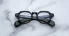 A pair of Jacques Marie Mage Kellerman black, round eyeglasses rests on a white marble surface, with brand details in white text visible on the inner arms through the clear lenses.