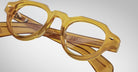 Close-up of Jacques Marie Mage Kellerman eyeglasses—round, translucent yellow frames with thick rims and folded arms—set against a light gray background.