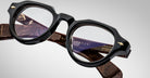 A close-up of Jacques Marie Mage Kellerman eyeglasses in black and brown shows thick frames, clear lenses, detailed hinges, gold accents, and engraved text on the arms.