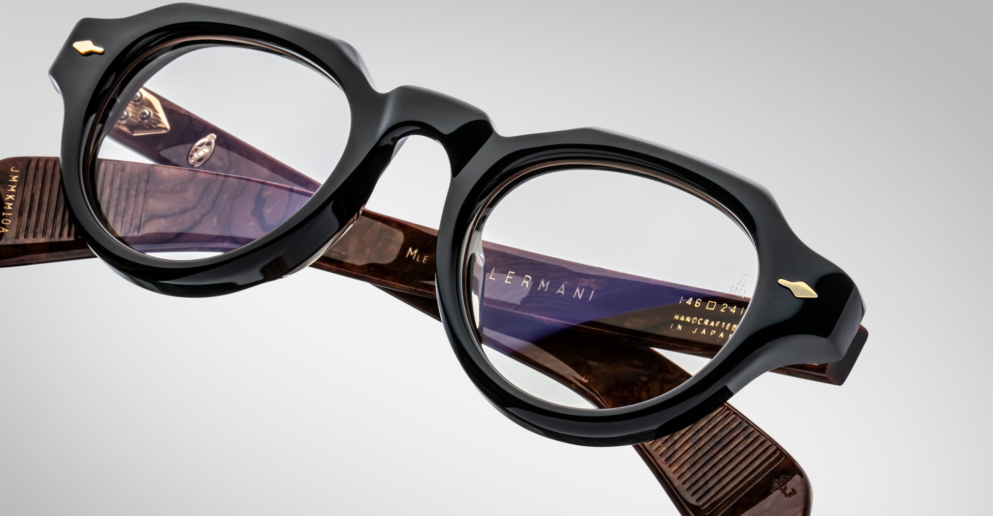 A close-up of Jacques Marie Mage Kellerman eyeglasses in black and brown shows thick frames, clear lenses, detailed hinges, gold accents, and engraved text on the arms.
