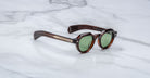 A pair of Jacques Marie Mage Kellerman sunglasses with round green lenses, thick dark brown tortoiseshell frames, and metallic arm accents rests on a white marble surface.