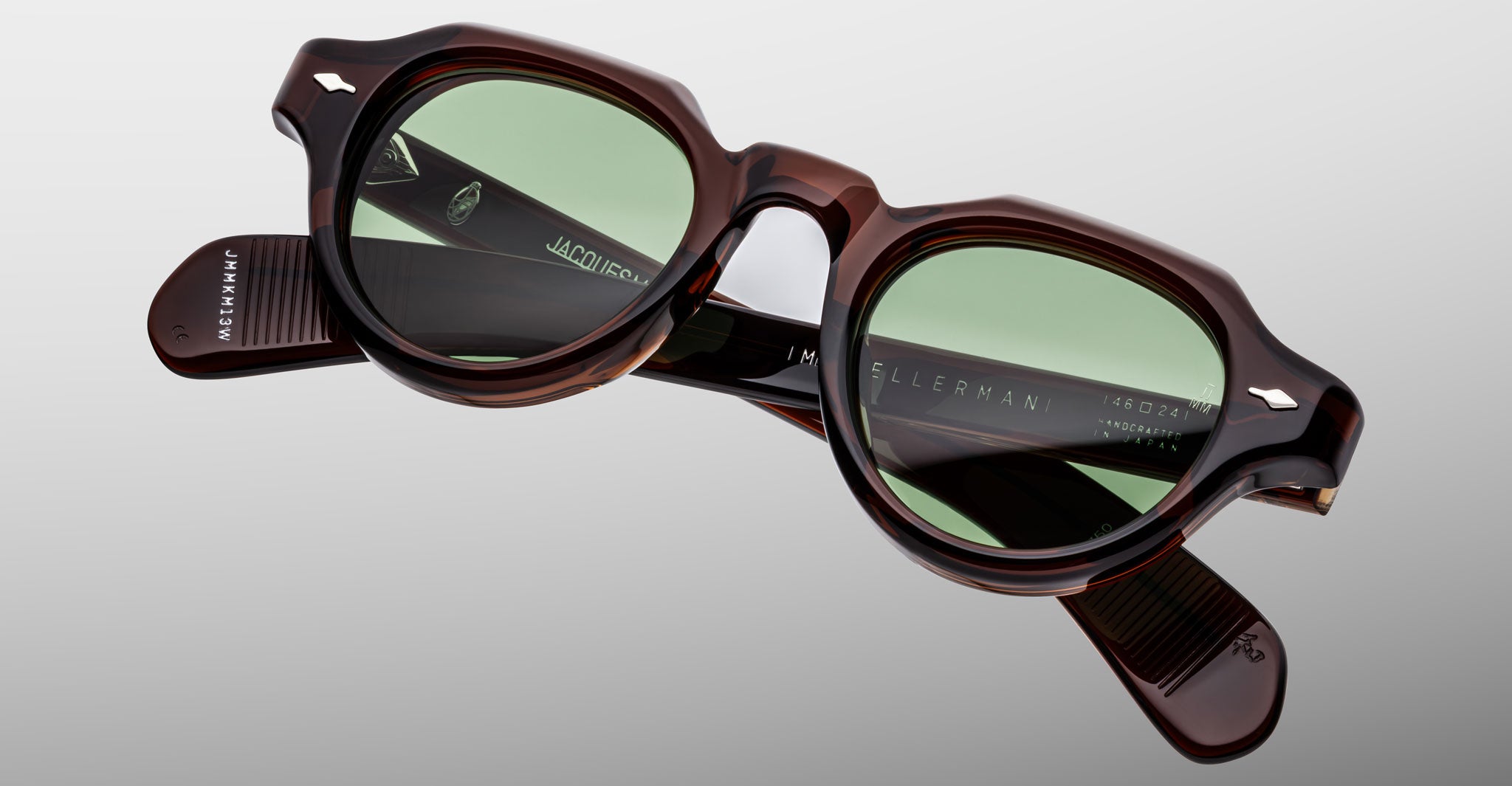 The Jacques Marie Mage Kellerman sunglasses feature round green-tinted lenses and thick tortoiseshell frames, displayed against a plain light gray background. The brand name is visible on the inside of the arms.