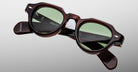 The Jacques Marie Mage Kellerman sunglasses feature round green-tinted lenses and thick tortoiseshell frames, displayed against a plain light gray background. The brand name is visible on the inside of the arms.