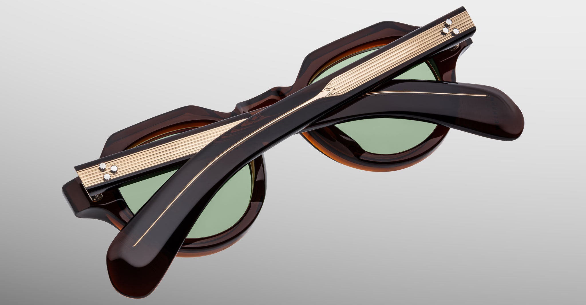 The Jacques Marie Mage Kellerman sunglasses, shown folded, feature round green lenses and dark brown frames with grooved metallic strips and small screw details on the arms, set against a light gray gradient background.