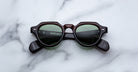 The Jacques Marie Mage Kellerman is a pair of black, thick-framed sunglasses with green tinted lenses displayed on a white marble surface.