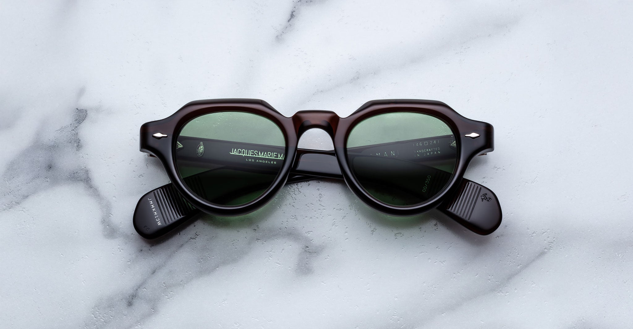 The Jacques Marie Mage Kellerman is a pair of black, thick-framed sunglasses with green tinted lenses displayed on a white marble surface.