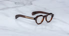 The Jacques Marie Mage Kellerman features dark brown, round eyeglasses with thick frames and metal temple accents, displayed on a white marble surface.
