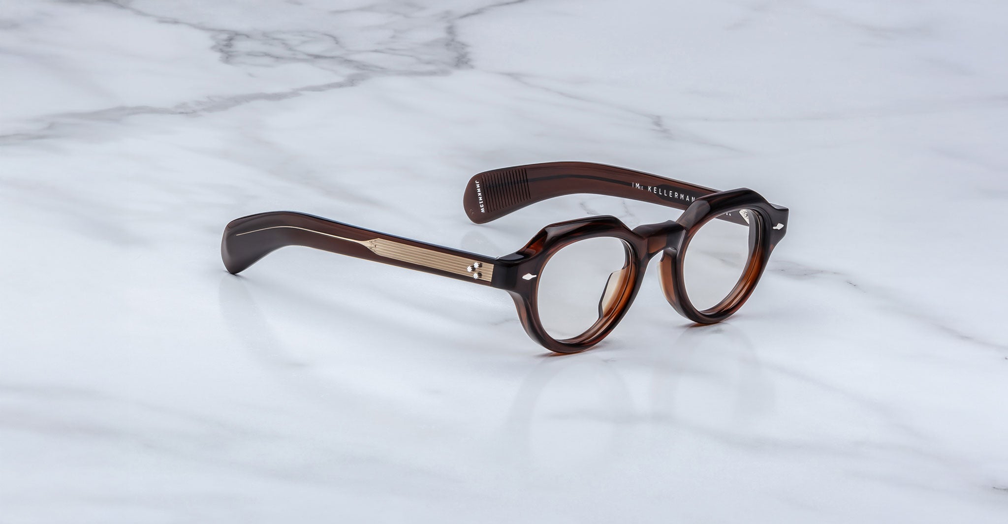The Jacques Marie Mage Kellerman features dark brown, round eyeglasses with thick frames and metal temple accents, displayed on a white marble surface.