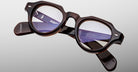 The Jacques Marie Mage Kellerman eyeglasses feature round, dark tortoiseshell thick frames with clear lenses, displayed on a light gray background. Branding and product details appear in white text on the inner arms.