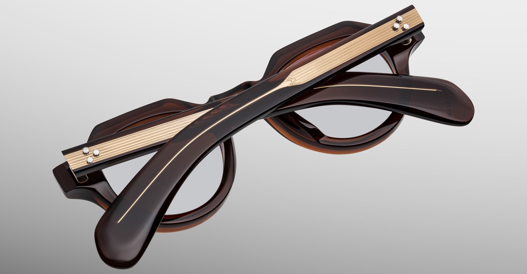 The Jacques Marie Mage Kellerman eyeglasses are shown folded, featuring thick dark brown frames with gold-toned metal temple accents against a matte gradient background.