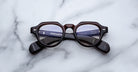 A pair of Jacques Marie Mage Kellerman round, dark-framed eyeglasses rests on a white marble surface, with text and part of a keyboard reflected in the lenses, hinting they face a computer screen.