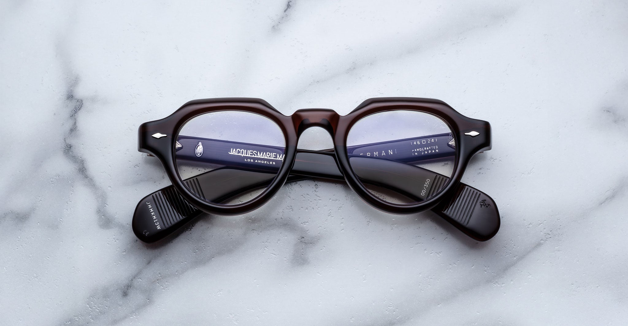 A pair of Jacques Marie Mage Kellerman round, dark-framed eyeglasses rests on a white marble surface, with text and part of a keyboard reflected in the lenses, hinting they face a computer screen.
