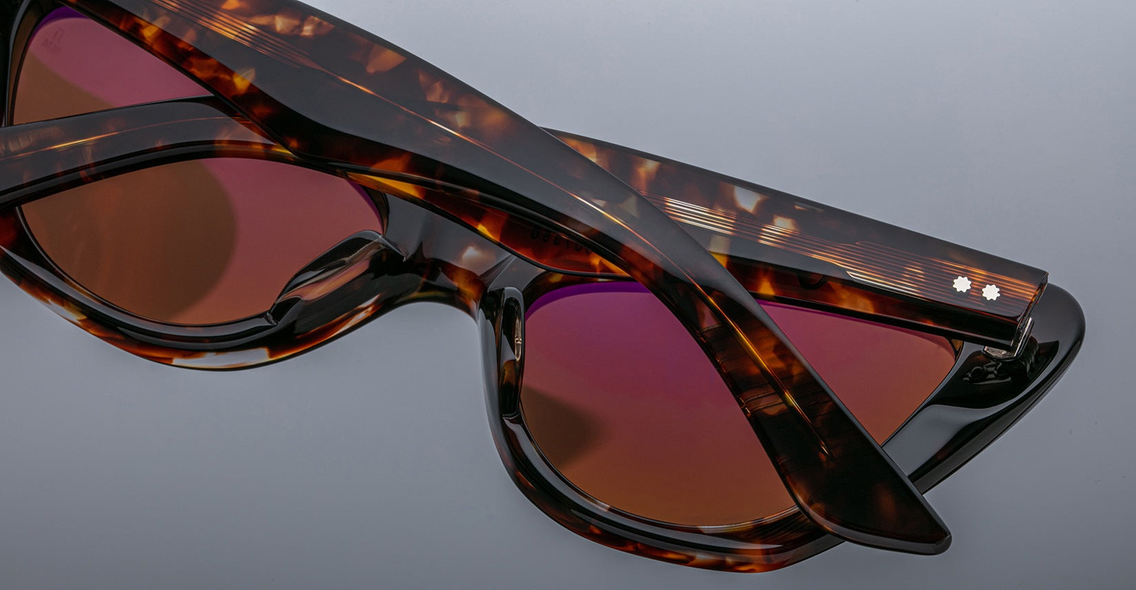 Close-up of two pairs of Jacques Marie Mage Kelly tortoiseshell sunglasses with dark reddish lenses, stacked on a reflective surface to highlight their distinctive frames and lens colors.