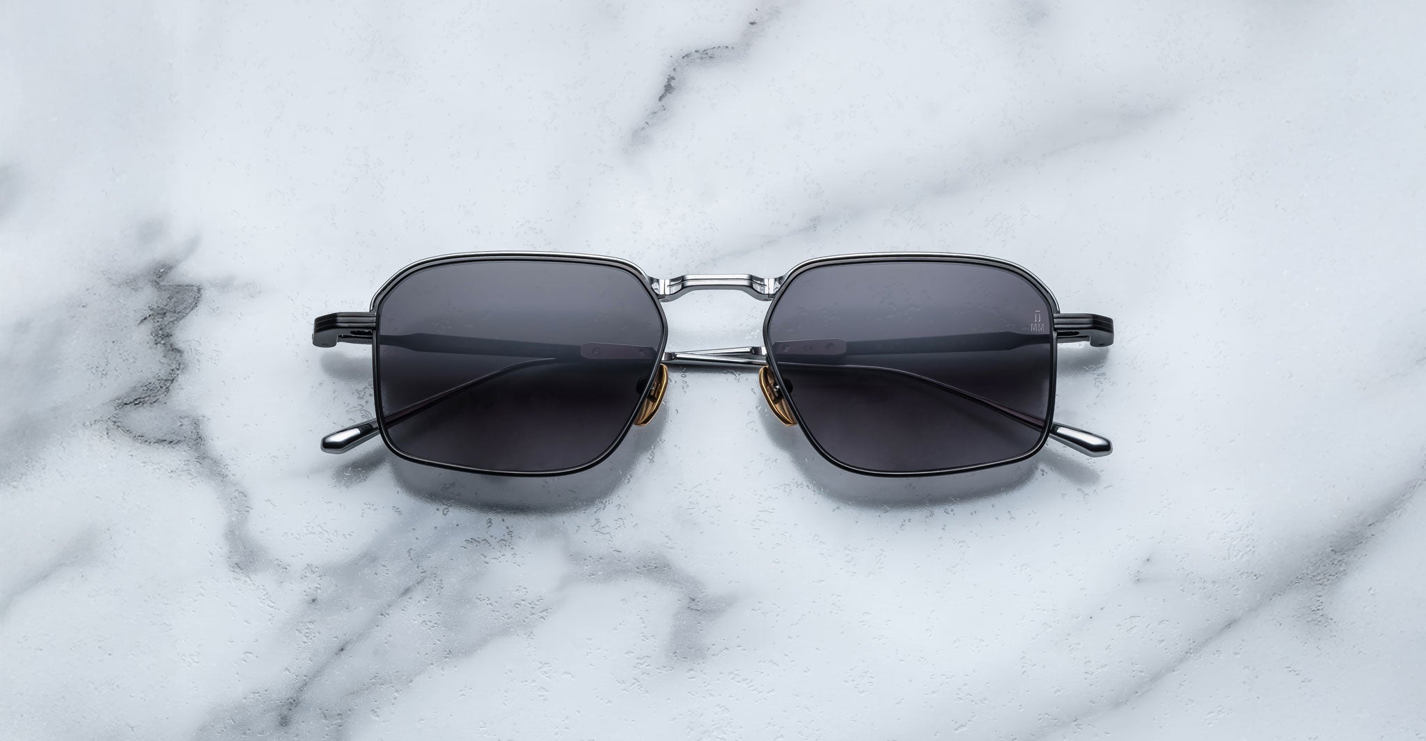 A pair of Jacques Marie Mage Koenig rectangular black sunglasses with metal frames and dark lenses rests on a white marble surface.