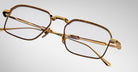 A close-up of Jacques Marie Mage Koenig gold-framed rectangular eyeglasses, featuring brown accents on the rims and temples, displayed against a plain light gray background.
