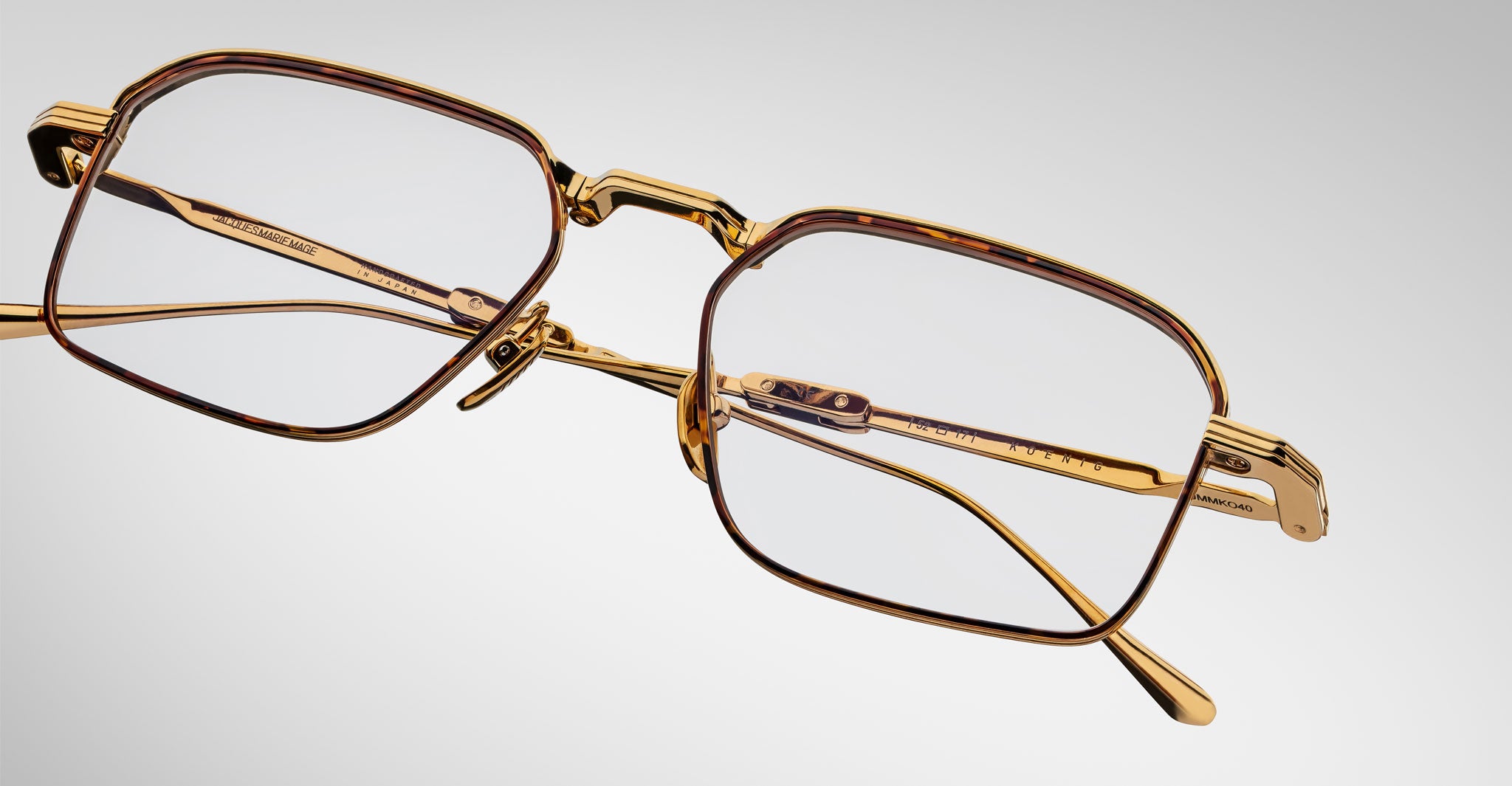 A close-up of Jacques Marie Mage Koenig gold-framed rectangular eyeglasses, featuring brown accents on the rims and temples, displayed against a plain light gray background.