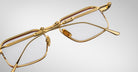 A close-up of Jacques Marie Mage Koenig gold-framed eyeglasses with clear lenses, set against a white background and angled to showcase the temple hinges and detailed design.
