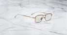 A pair of Jacques Marie Mage Koenig gold, square-framed eyeglasses with clear lenses rests on a white marble surface.
