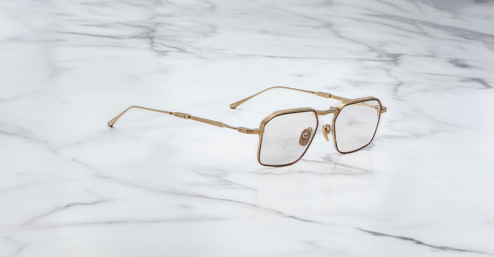 A pair of Jacques Marie Mage Koenig gold, square-framed eyeglasses with clear lenses rests on a white marble surface.
