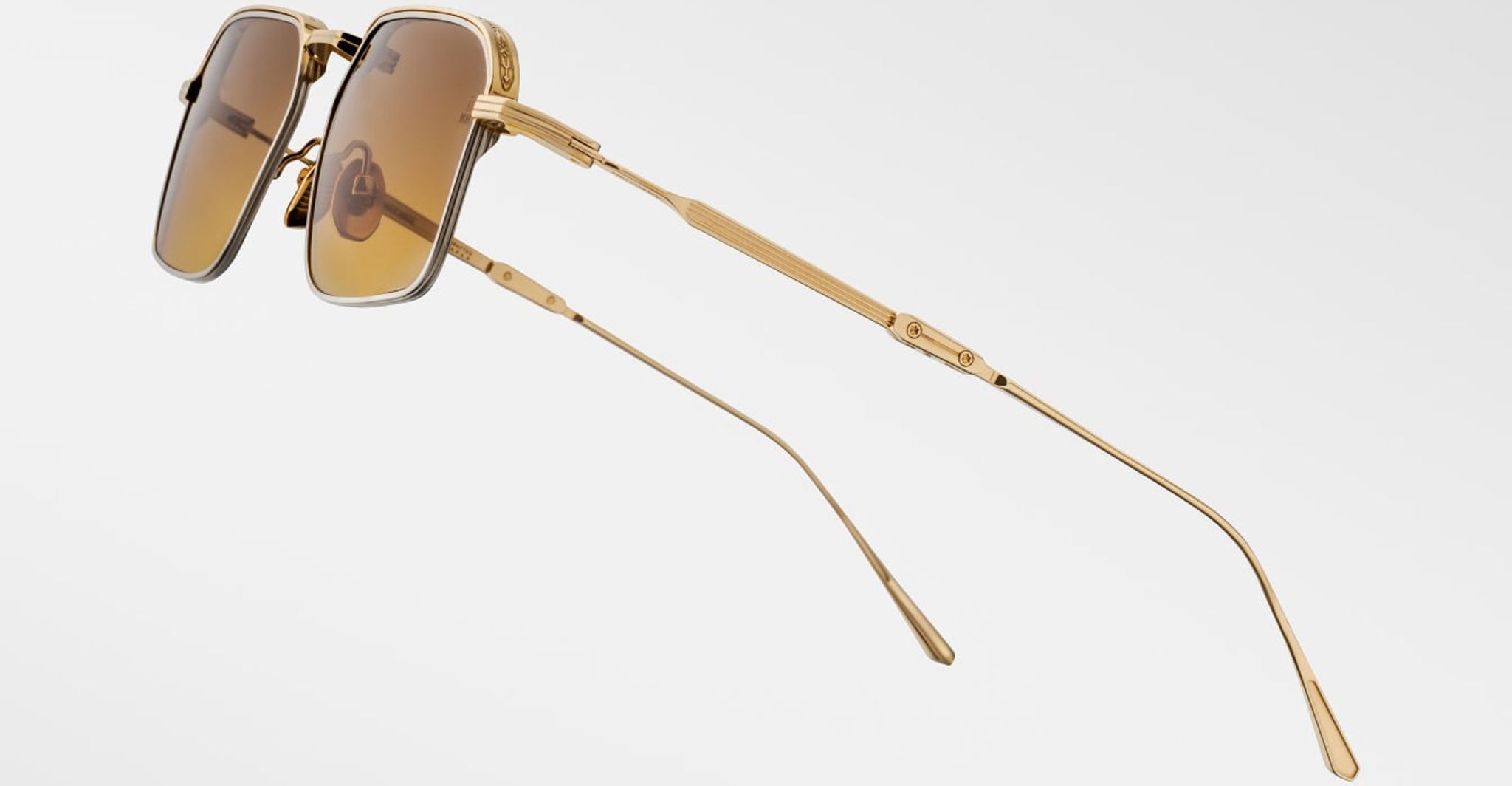 The Jacques Marie Mage Koenig sunglasses feature gold frames and brown-tinted square lenses, displayed at an angle on a plain light background. Slim temples and arms showcase a refined textured design.