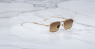 A pair of Jacques Marie Mage Koenig gold-rimmed, square-shaped sunglasses with brown gradient lenses rests on a white marble surface with gray veining.