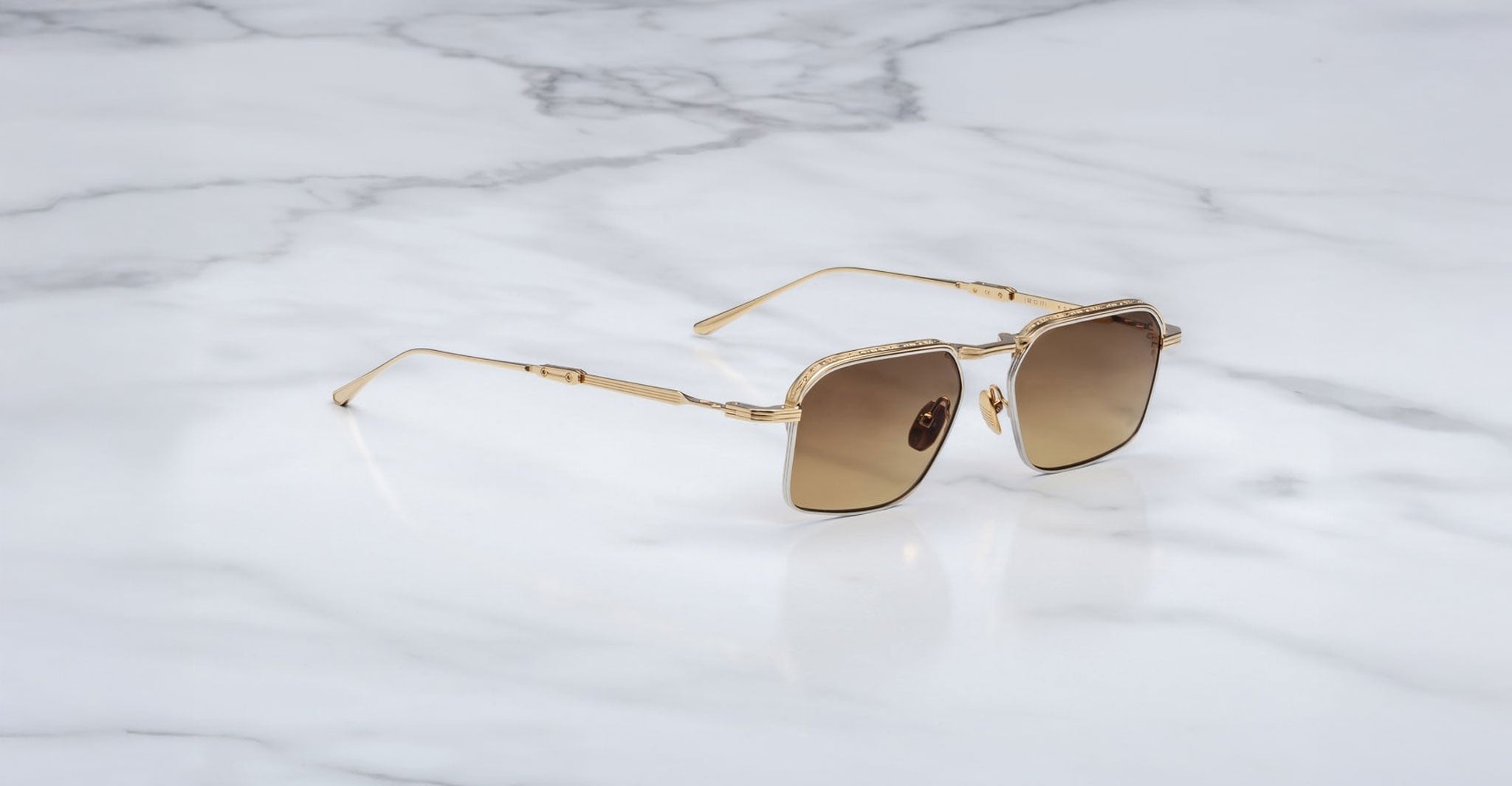 A pair of Jacques Marie Mage Koenig gold-rimmed, square-shaped sunglasses with brown gradient lenses rests on a white marble surface with gray veining.