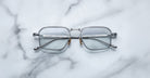 A pair of Jacques Marie Mage Koenig rectangular silver metal eyeglasses with clear lenses is displayed open on a white marble surface, temples facing upward.