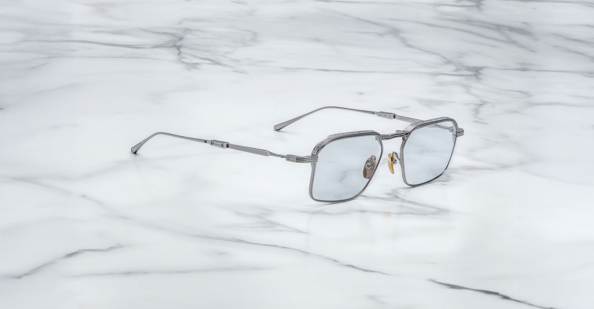 Jacques Marie Mage Koenig silver metal-framed square sunglasses with light-tinted lenses rest on a white marble surface.