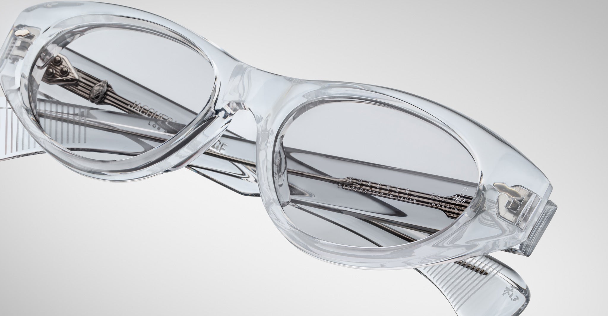 The Jacques Marie Mage Krasner eyeglasses, featuring clear, oval-shaped lenses with transparent frames and silver accents, are shown close up at an angle against a light gray background to highlight their design.