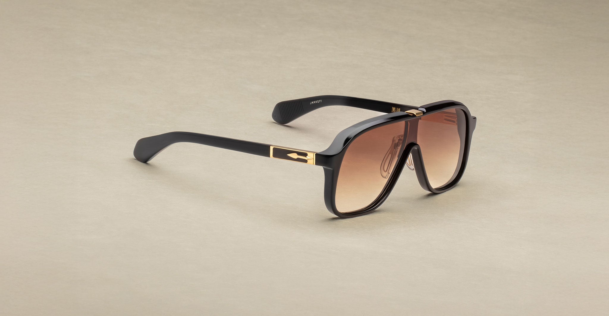 The Jacques Marie Mage Kurokawa sunglasses feature an oversized black frame, brown gradient lenses, and gold temple accents, displayed on a light beige surface.