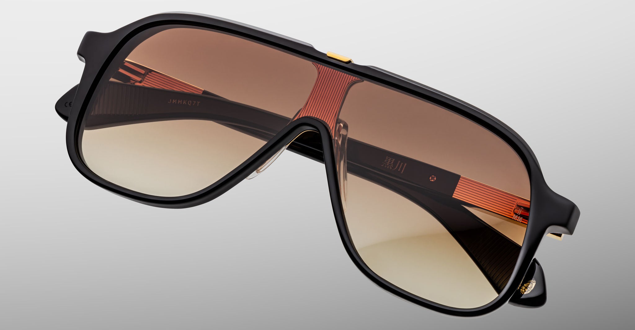 The Jacques Marie Mage Kurokawa sunglasses feature stylish oversized black frames, brown gradient lenses, gold accents, and subtle orange details on the arms, shown against a light gray background.