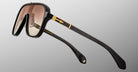 Close-up of Jacques Marie Mage Kurokawa sunglasses featuring black frames, gold accents, and brown gradient lenses, displayed at an angle against a plain gray background.