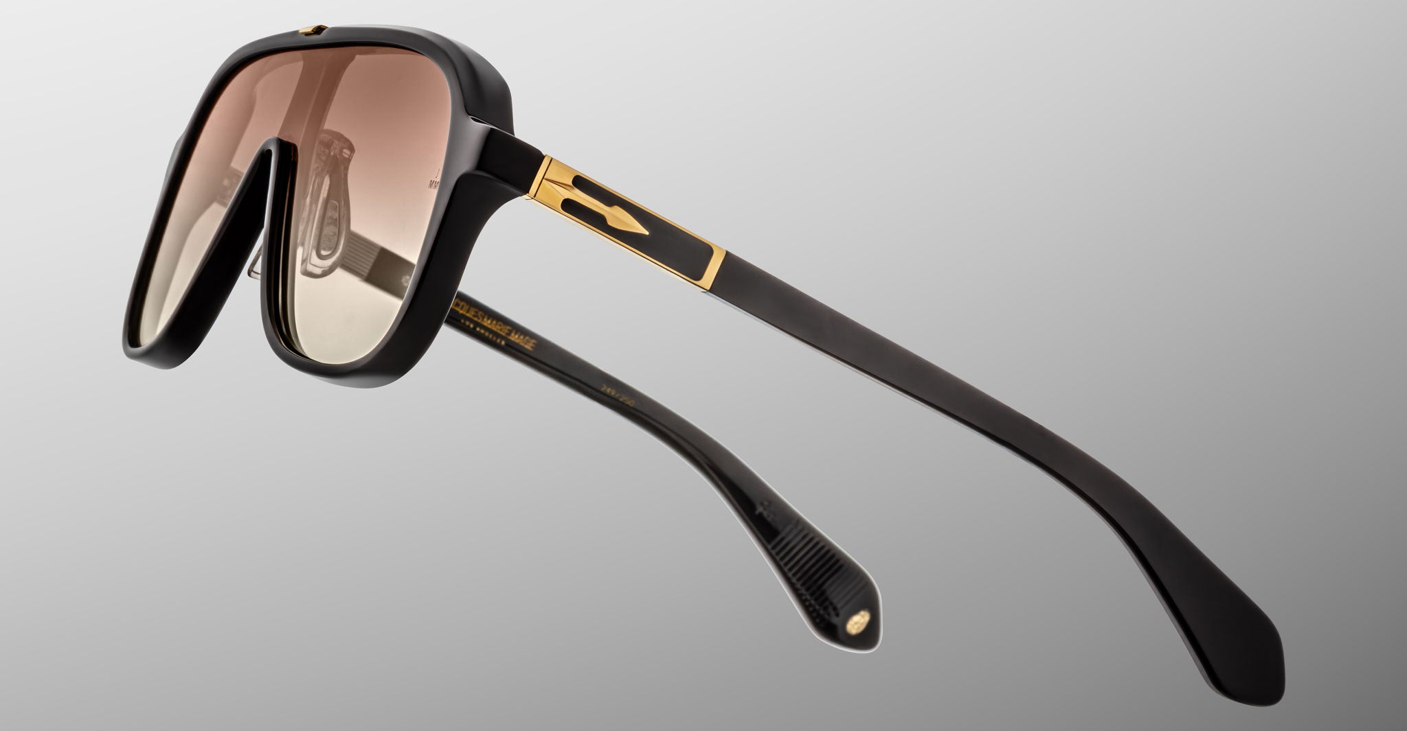 Close-up of Jacques Marie Mage Kurokawa sunglasses featuring black frames, gold accents, and brown gradient lenses, displayed at an angle against a plain gray background.