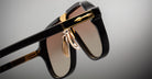 Close-up of Jacques Marie Mage Kurokawa luxury black sunglasses with gold accents and brown gradient lenses, highlighting the detailed hinges and nose pads against a neutral background.