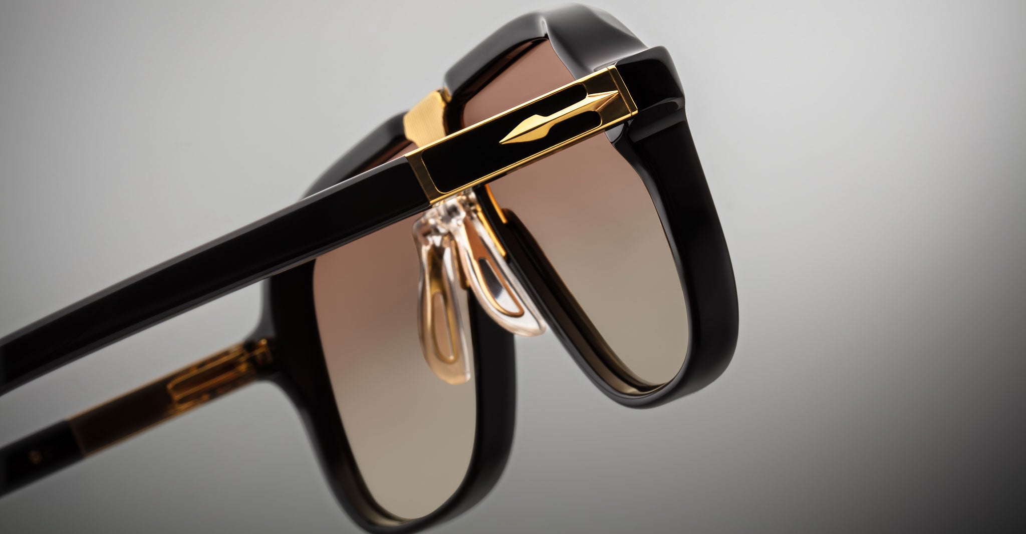 Close-up of Jacques Marie Mage Kurokawa luxury black sunglasses with gold accents and brown gradient lenses, highlighting the detailed hinges and nose pads against a neutral background.