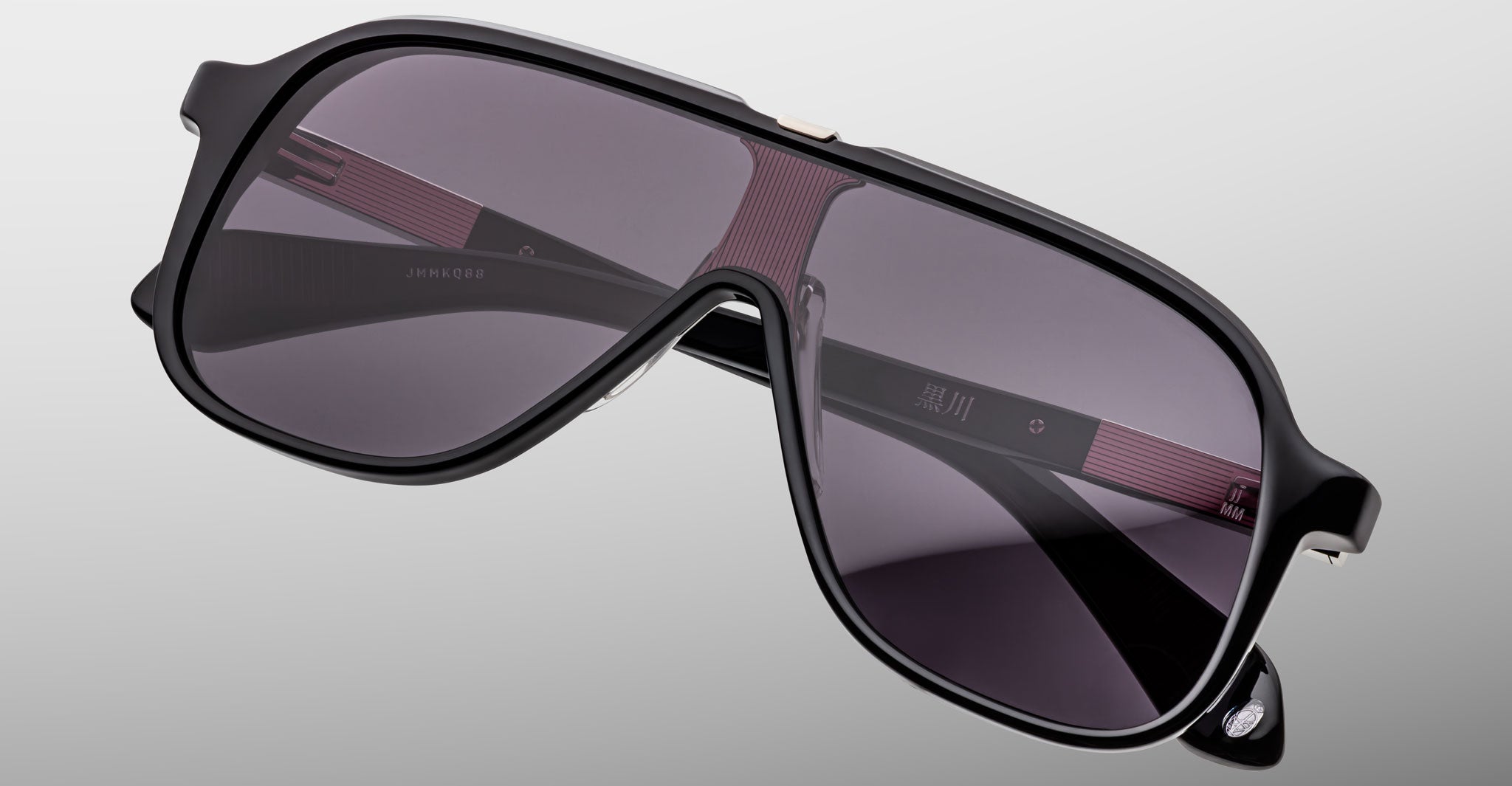 Close-up of the Jacques Marie Mage Kurokawa: stylish oversized black sunglasses with dark tinted lenses and thick frames, set against a plain gray background. The temples feature red accents and subtle white branding details.