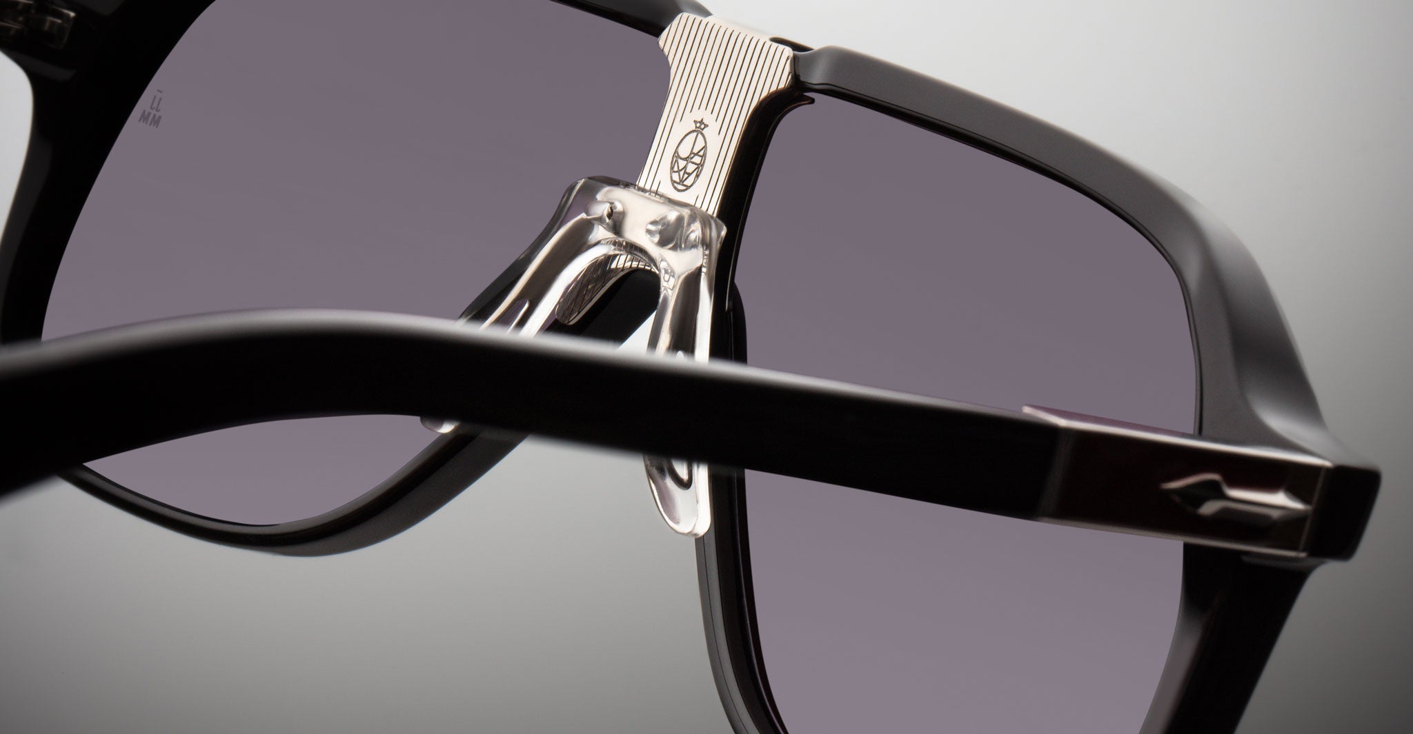 Close-up of Jacques Marie Mage Kurokawa black sunglasses with a silver metal nose bridge, clear nose pads, and a small engraved logo on the bridge and one arm, set against a neutral background.