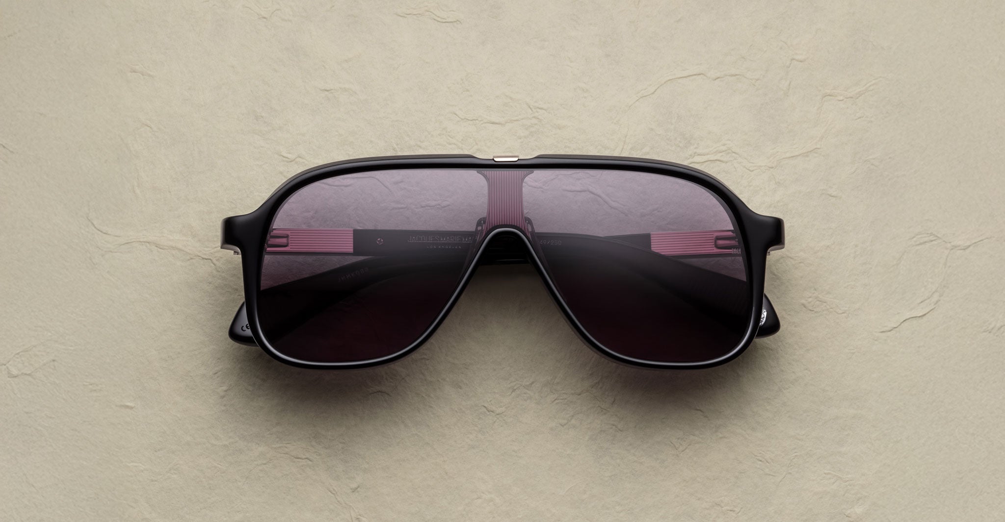 The Jacques Marie Mage Kurokawa sunglasses, featuring a large black aviator design with dark lenses, rest on a beige textured surface.