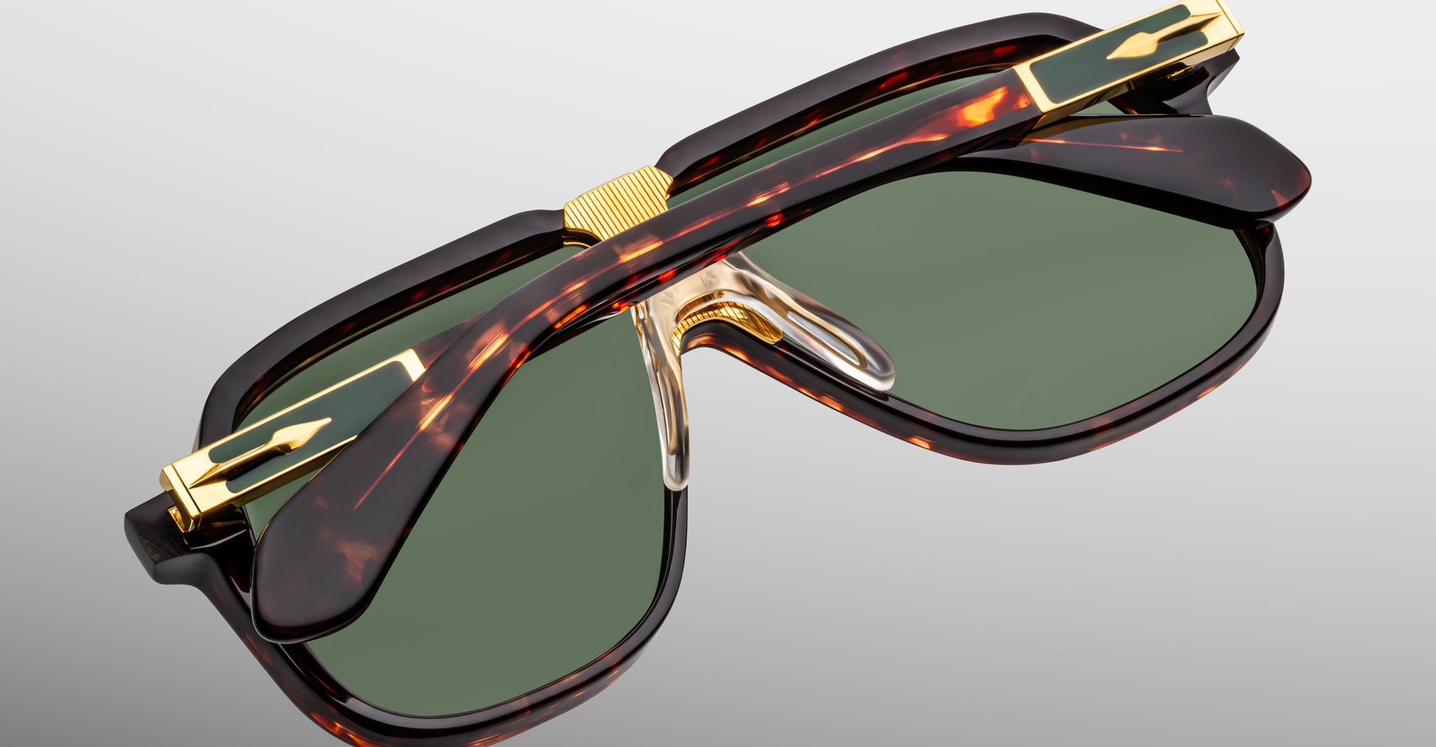 Close-up of Jacques Marie Mage Kurokawa sunglasses featuring green lenses, tortoiseshell frames, and gold accents, showcasing the unique bridge and detailed hinge design against a light gray background.