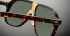 Close-up of Jacques Marie Mage Kurokawa sunglasses with tortoiseshell frames, green-tinted lenses, gold nose bridge detailing, and transparent nose pads against a plain gray background.