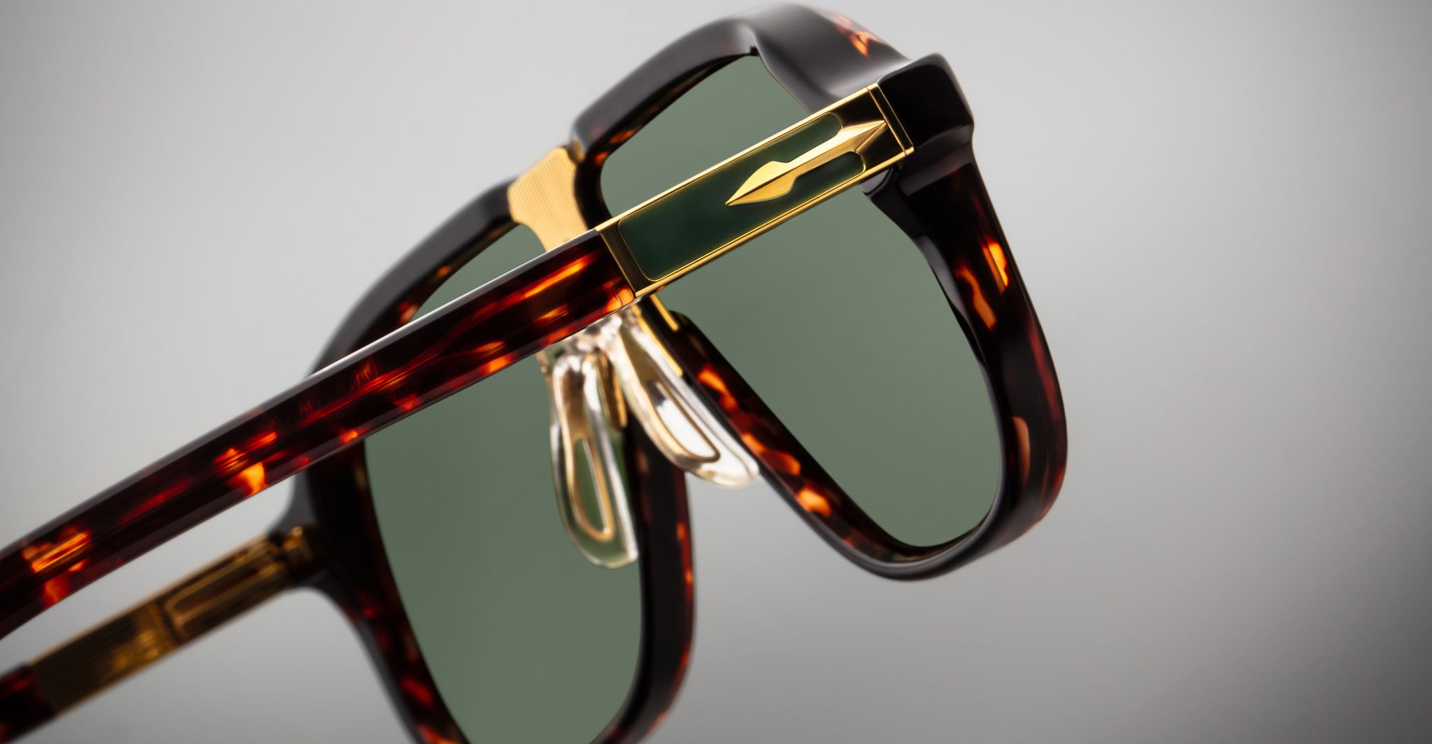 Close-up of Jacques Marie Mage Kurokawa sunglasses featuring tortoiseshell frames, green lenses, gold accents, and adjustable nose pads, set against a plain gray background.