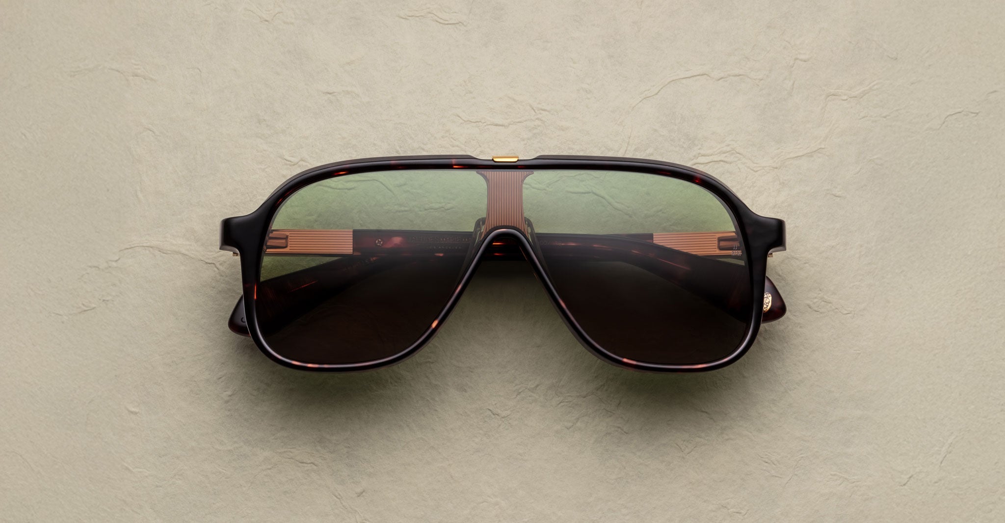 The Jacques Marie Mage Kurokawa sunglasses, featuring large dark lenses and a tortoiseshell frame, rest on a textured beige surface with reflections of stripes and light in the lenses.